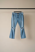 Light blue distressed denim jeans with ripped details on the thighs, hanging on a wooden hanger against a gray fabric backdrop.