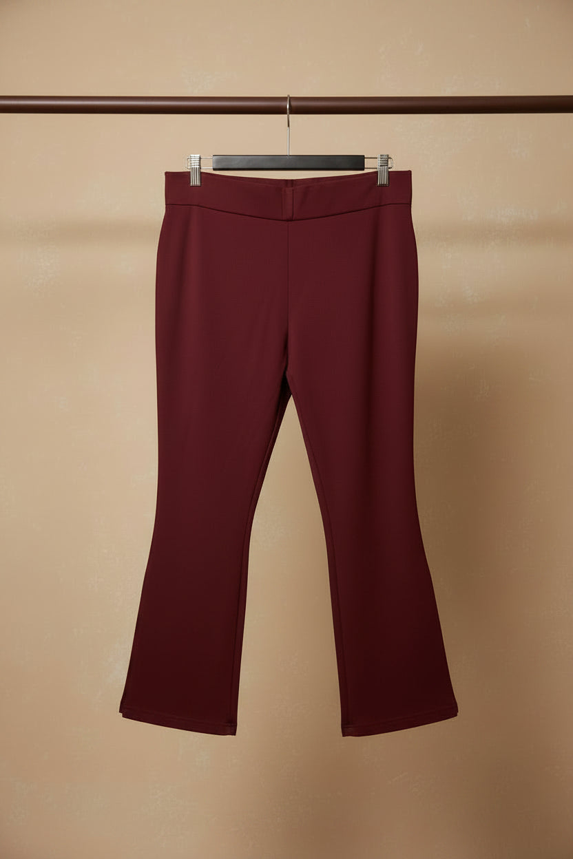 A pair of burgundy cropped pants with side slits, hanging on a wooden hanger against a wrinkled white fabric backdrop.
