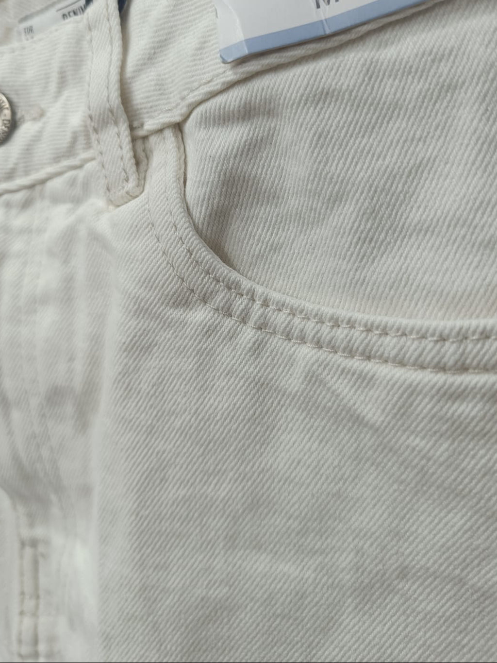 Close-up of a pair of light beige or off-white denim jeans, showing the waistband, button, pocket seam, and a size tag marked M / 28.