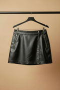 A black faux-leather mini skirt with two silver zipper details on the sides, hanging on a wooden hanger against a light beige fabric background.