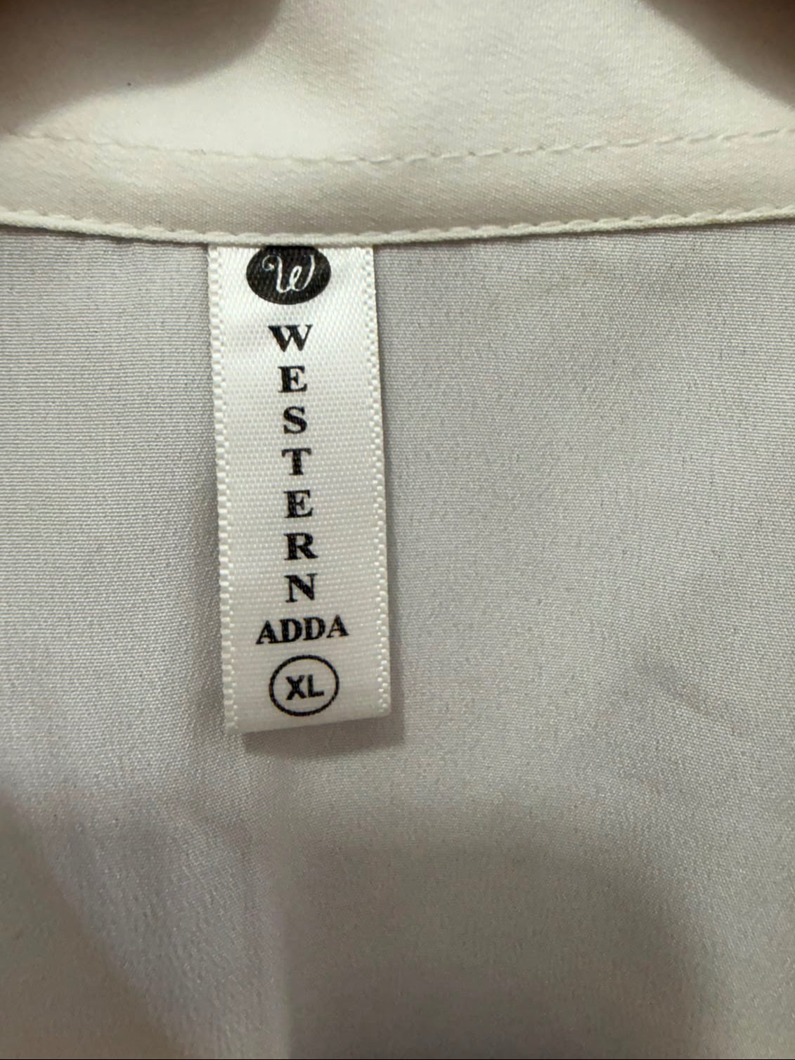 Close-up of a garment's interior neck label reading 'WESTERN ADDA' with a small 'W' logo and size 'XL' in black text on a white fabric tag.
