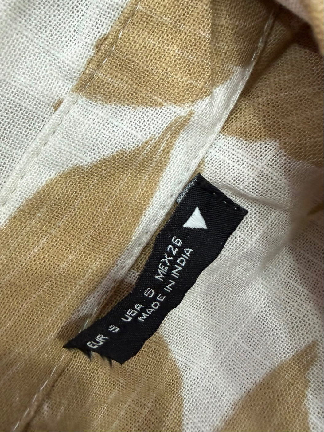 Close-up of a beige and off-white patterned fabric with a black sewn-in label reading 'BURS S. LEE S. MEX-256 MADE IN INDIA'.