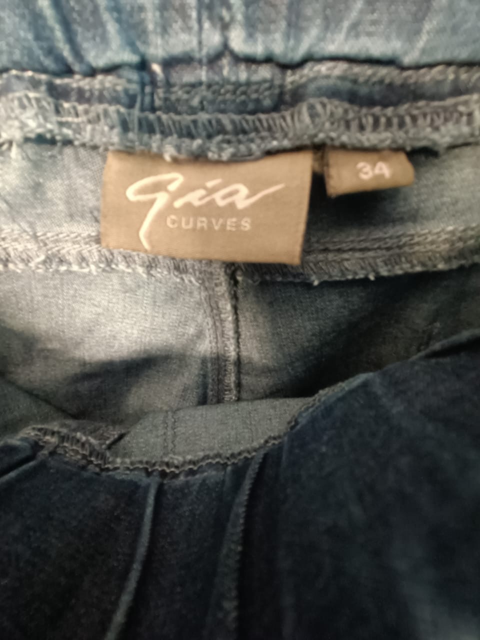 Close-up of the waistband of blue denim jeans showing a tan brand label that reads 'Gia Curves' and size '34'.
