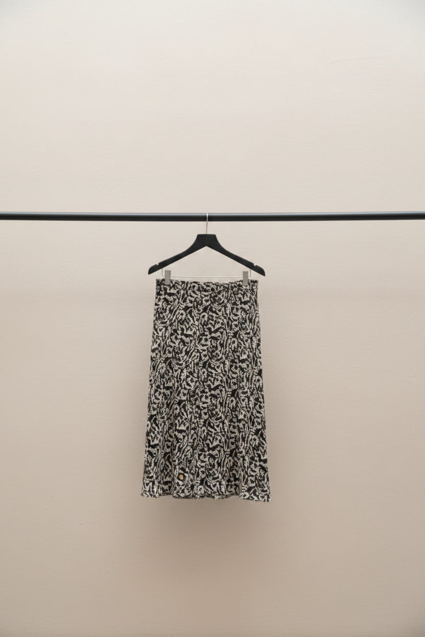 A black and white patterned midi skirt hanging from a wooden clothes hanger on a tree branch in an outdoor patio setting.