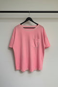 A pink short-sleeve crewneck t-shirt with a chest pocket, hanging on a wooden hanger against a wrinkled white fabric backdrop.