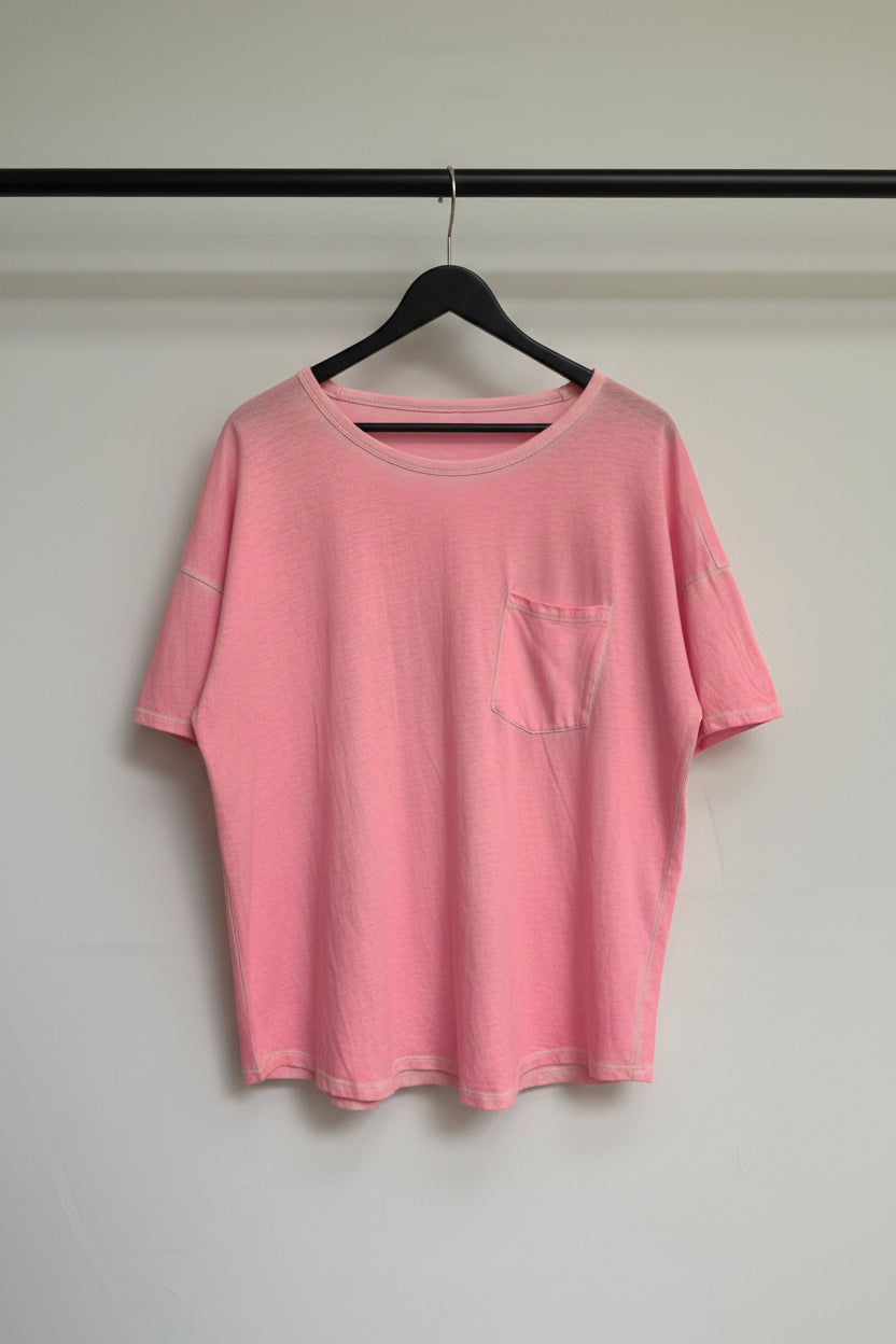 A pink short-sleeve crewneck t-shirt with a chest pocket, hanging on a wooden hanger against a wrinkled white fabric backdrop.