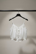 A white camisole-style top with lace trim and a button front, hanging outdoors on tree branches with green leaves and a building visible in the background.