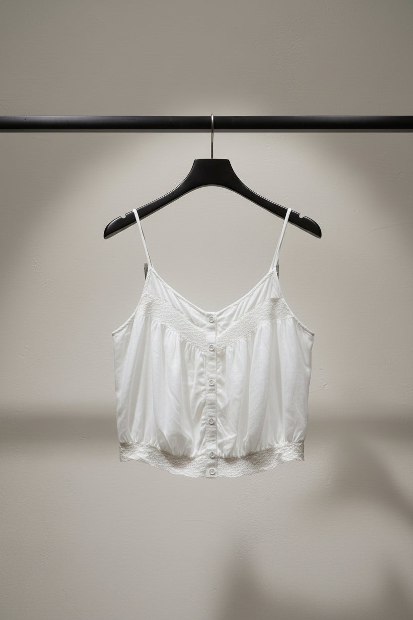 A white camisole-style top with lace trim and a button front, hanging outdoors on tree branches with green leaves and a building visible in the background.