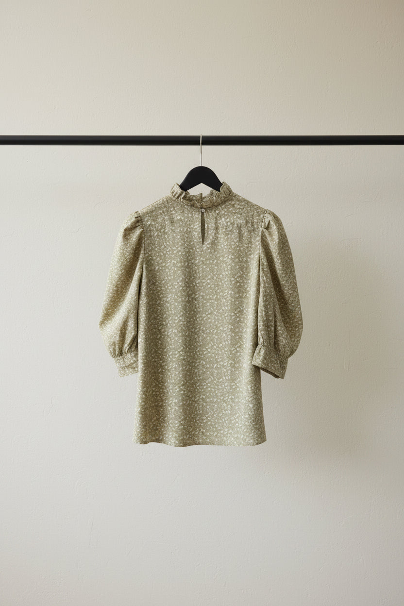 A beige and white floral print blouse with a high collar and puffed sleeves, hanging on a wooden hanger against a dark red background.