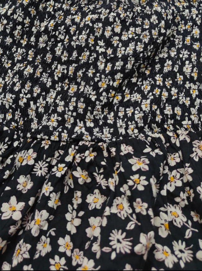 A close-up of a black smocked dress with a dense white and yellow daisy floral print, featuring thin spaghetti straps.