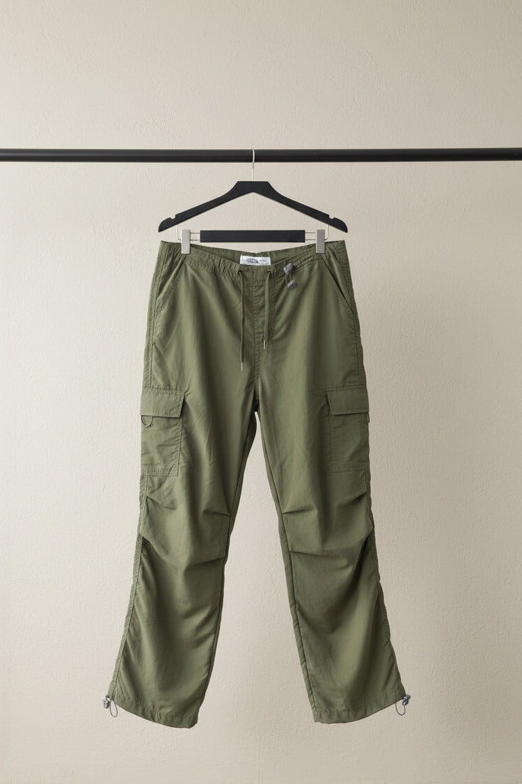 Olive green cargo pants with side flap pockets and elasticized cuffs, hanging against a gray textured backdrop.