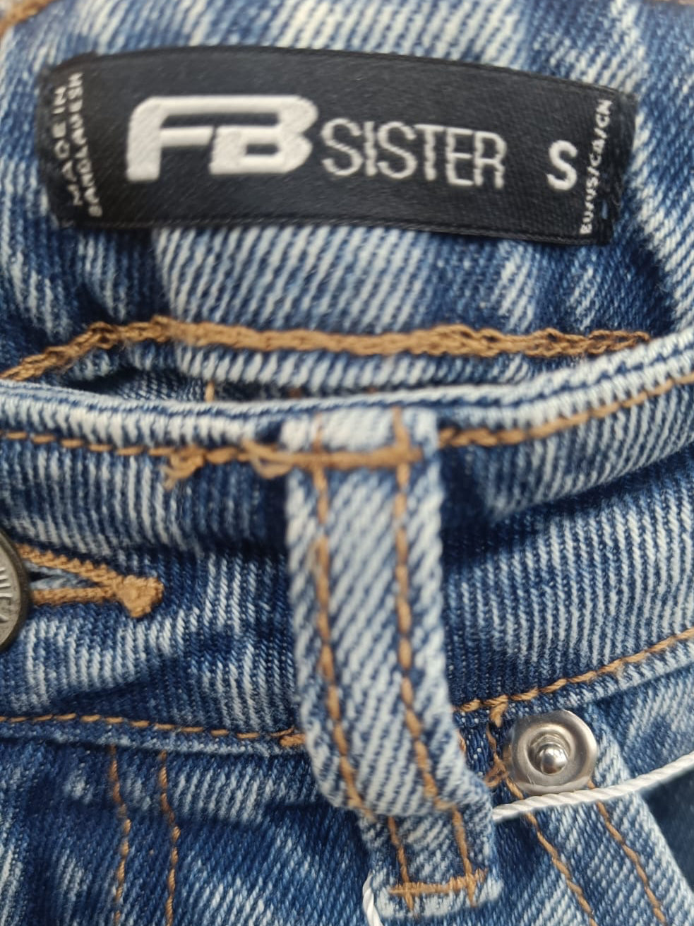 Close-up of a pair of blue denim jeans showing the waistband, belt loop, button, and a black brand label that reads FB SISTER.