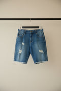 Light blue distressed denim shorts with frayed hems and ripped details, hanging on a wooden hanger against a red background.