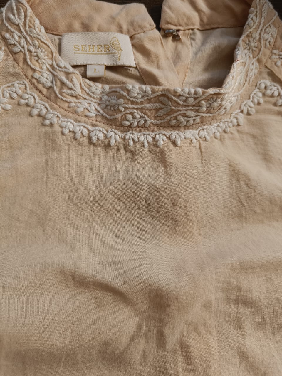 Close-up of a beige linen top with intricate white floral embroidery around the round neckline. The brand label 'SEHER' is visible inside the collar.