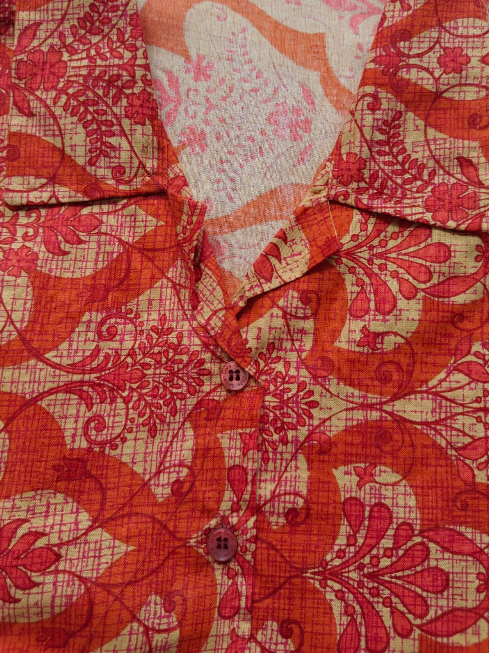 A close-up of a collared button-front shirt with an ornate floral and scroll pattern in red and orange over a beige background.