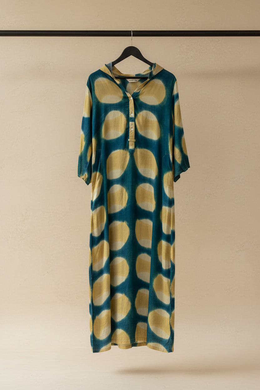 A long-sleeve tunic dress with a teal blue and yellow tie-dye pattern, hanging on a wooden hanger against an outdoor patio backdrop.