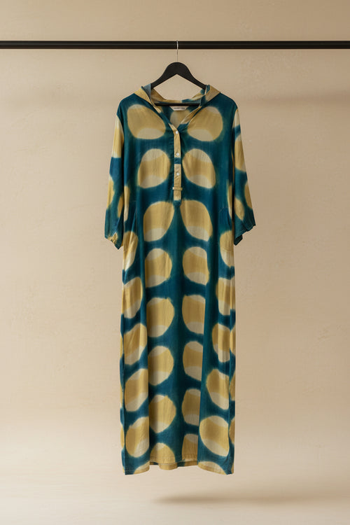 A long-sleeve tunic dress with a teal blue and yellow tie-dye pattern, hanging on a wooden hanger against an outdoor patio backdrop.