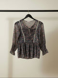 A women's long-sleeve blouse with a dark floral pattern, featuring white lace trim along the split neckline and ruffled cuffs, laid flat on a white fabric background.