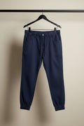 Dark navy blue tapered pants with a drawstring waist and elastic cuffs, hanging on a wooden hanger against a white fabric backdrop.