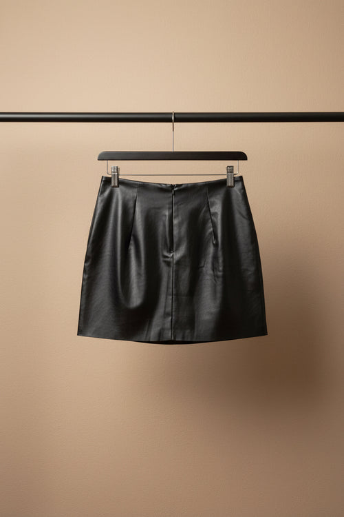 A black faux-leather mini skirt with a front zipper, hanging from a wooden hanger with metal clips against a light-colored fabric background.