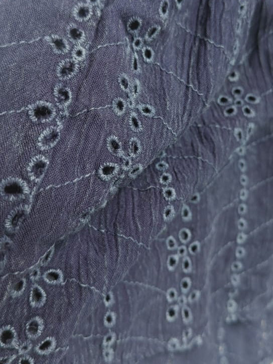 Close-up of a textured, eyelet embroidered fabric in a muted slate blue or grayish purple color, draped against a plain off-white background.
