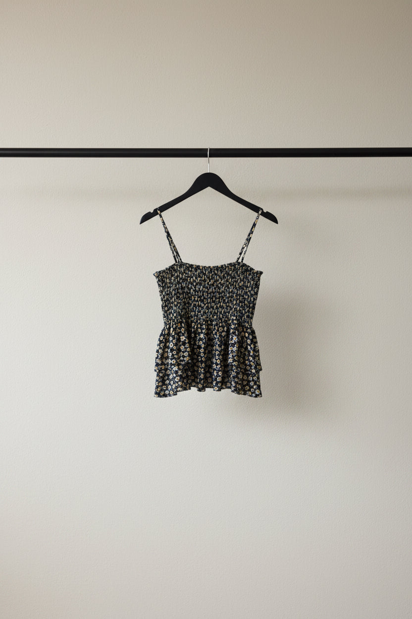 A sleeveless black camisole top with a dense small floral print in white and beige, featuring thin shoulder straps and a peplum-style ruffled hem, laid flat on a white fabric background.