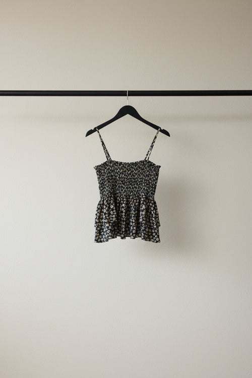 A sleeveless black camisole top with a dense small floral print in white and beige, featuring thin shoulder straps and a peplum-style ruffled hem, laid flat on a white fabric background.