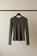 A dark charcoal gray long-sleeve crewneck top hanging on a wooden hanger outdoors against a background of tree branches and leaves.