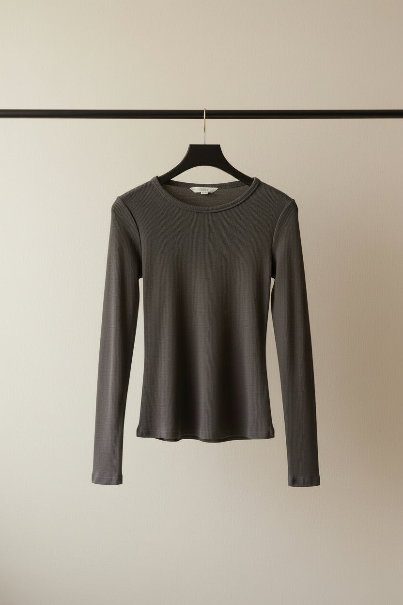 A dark charcoal gray long-sleeve crewneck top hanging on a wooden hanger outdoors against a background of tree branches and leaves.