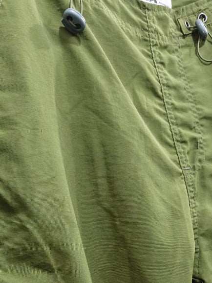 Olive green cargo pants with side flap pockets and metal D-ring hardware, shown hanging against a light gray fabric backdrop.