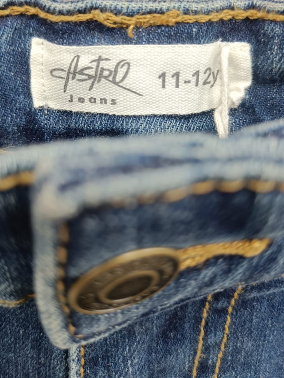 Close-up of a pair of blue denim jeans showing the inner waistband label which reads 'Astró Jeans 11-12y', along with yellow contrast stitching and a brass button.