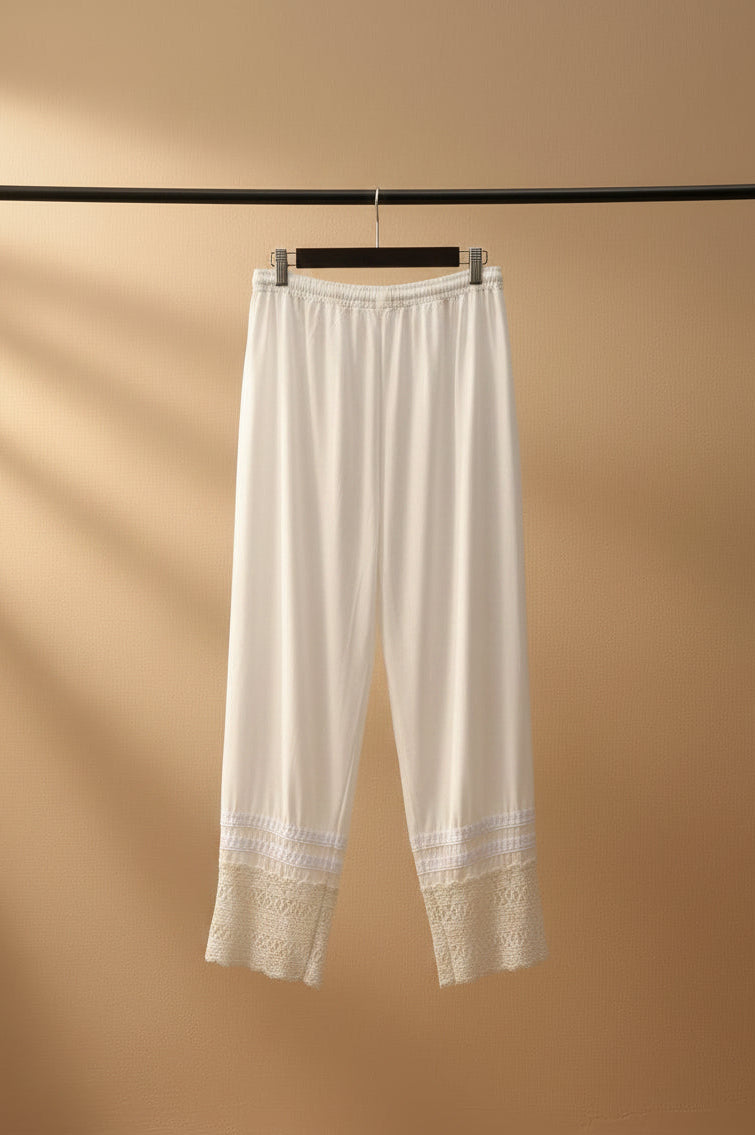 White cropped pants with lace hem detailing, hanging from a wooden hanger outdoors against a backdrop of tree branches and a tiled patio.