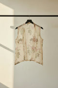 A sleeveless, open-front sheer vest with a pink and green floral print, hanging on a wooden hanger against a dark brown wooden slat wall.