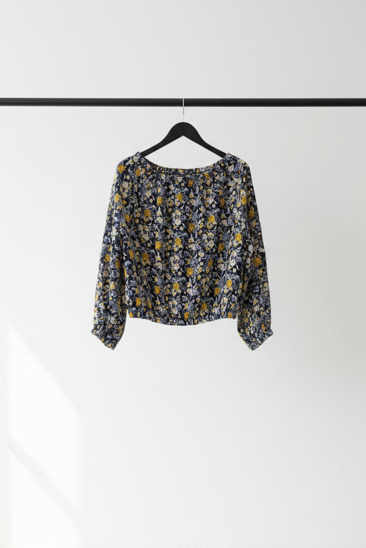A long-sleeve off-the-shoulder blouse with a dense yellow, white, and light blue floral print on a dark navy background, hanging on a wooden hanger from tree branches.
