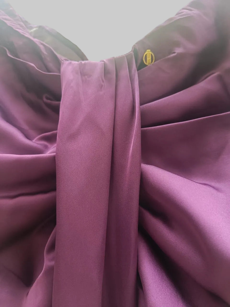 A close-up of draped purple fabric with gathered folds and a small gold metal grommet near the top edge, with blurred green leaves visible in the background.