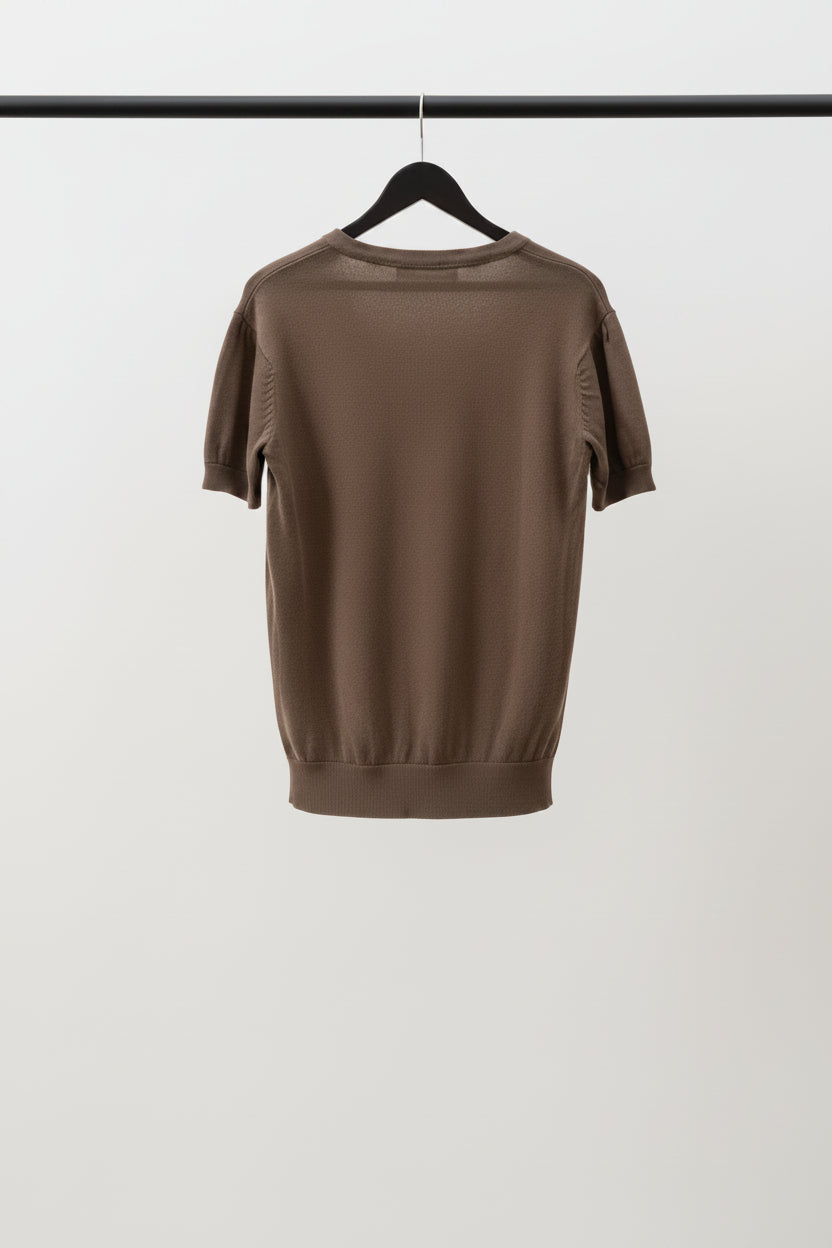 A brown short-sleeve knit top hanging on a hanger, showing the back view with a faded heart graphic on the upper back, set against a background of potted plants.