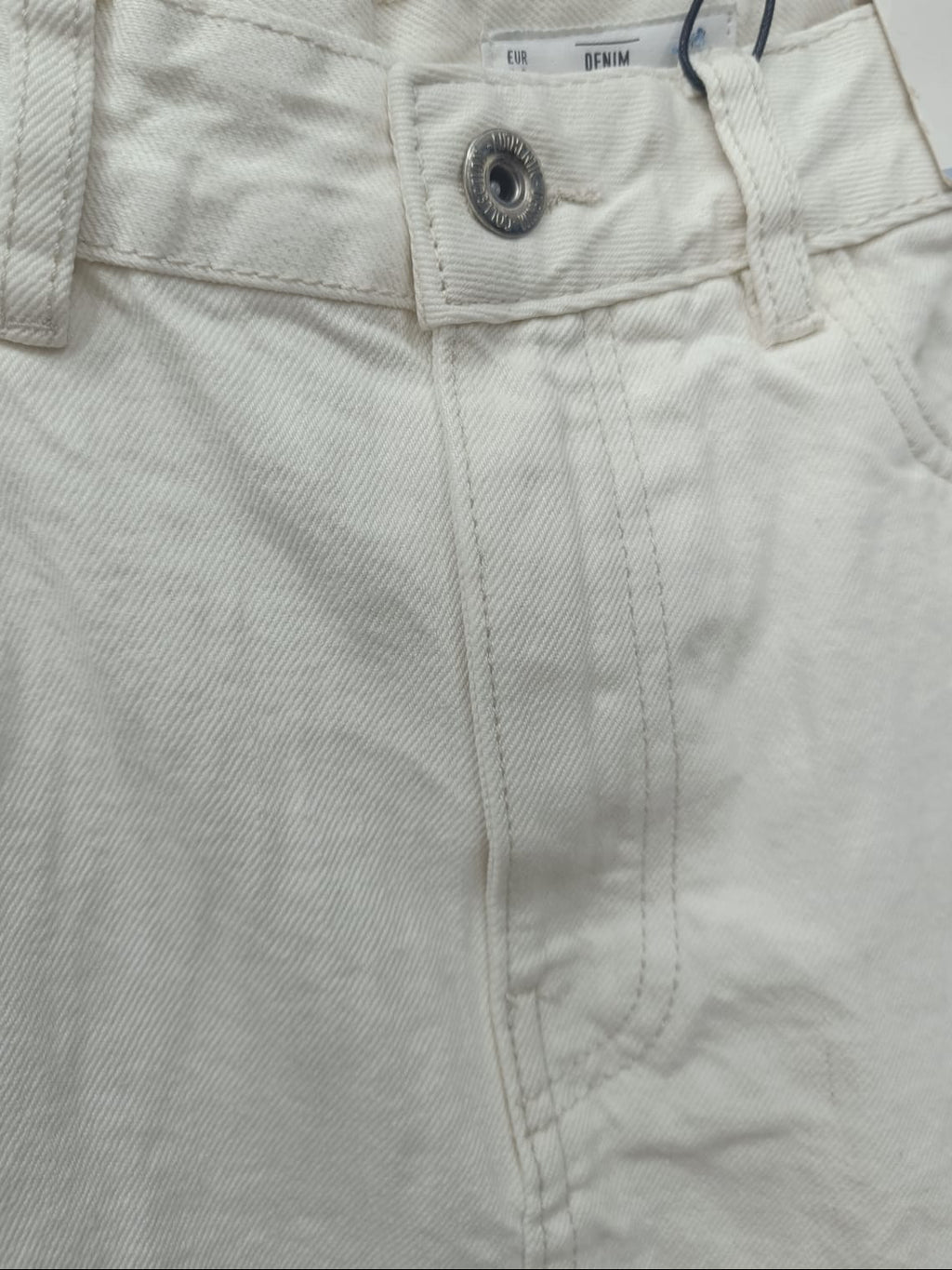 Close-up of the waistband of off-white denim jeans, showing the button closure, belt loops, and stitching details.
