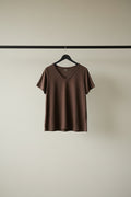 A brown short-sleeve V-neck t-shirt hanging on a wooden hanger outdoors against a background of green trees.
