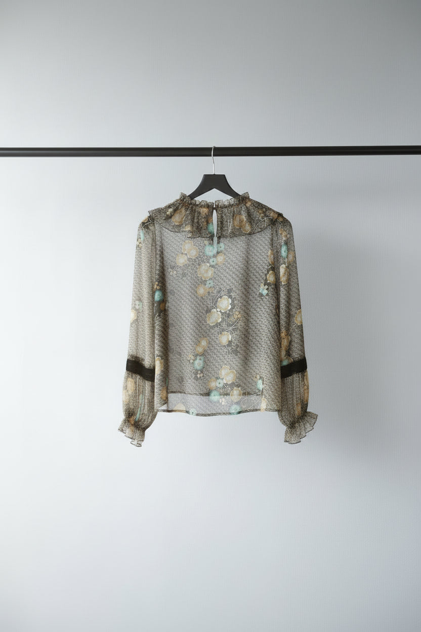 A long-sleeve blouse with a dense abstract floral pattern in yellow, blue, and green on a black background, laid flat on a white fabric surface.