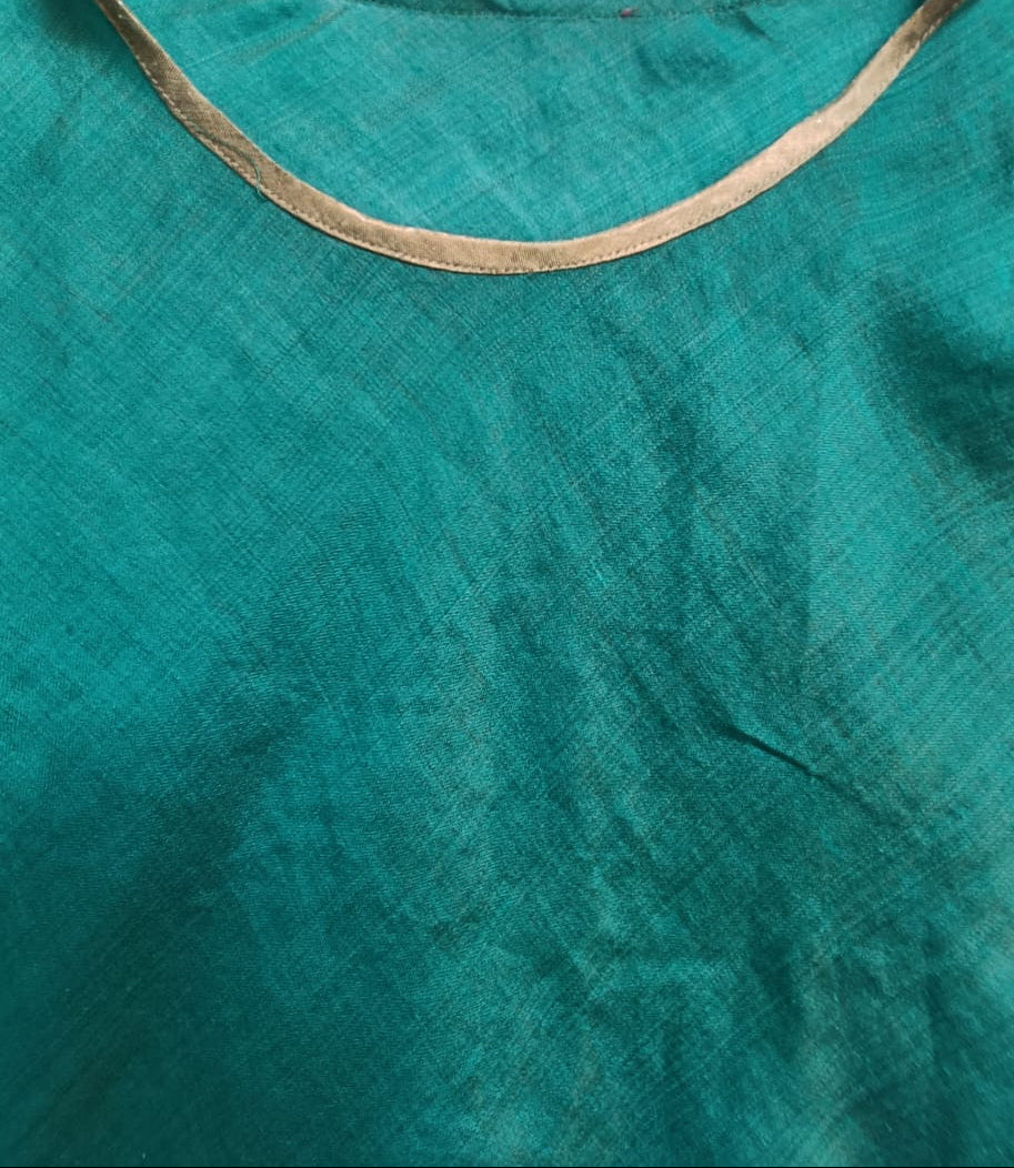 A close-up of a green textured fabric garment, likely a kurta or blouse, featuring a contrasting beige/tan embroidered or printed border around the scoop neckline.