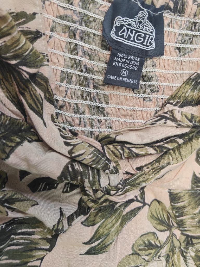 Close-up of a sleeveless top or dress neckline with a tropical leaf print in beige, green, and brown tones, featuring a black fabric brand label reading ANGII.