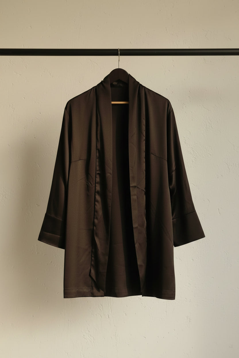 A black open-front kimono-style robe or cardigan hanging on a wooden hanger against a white fabric backdrop.