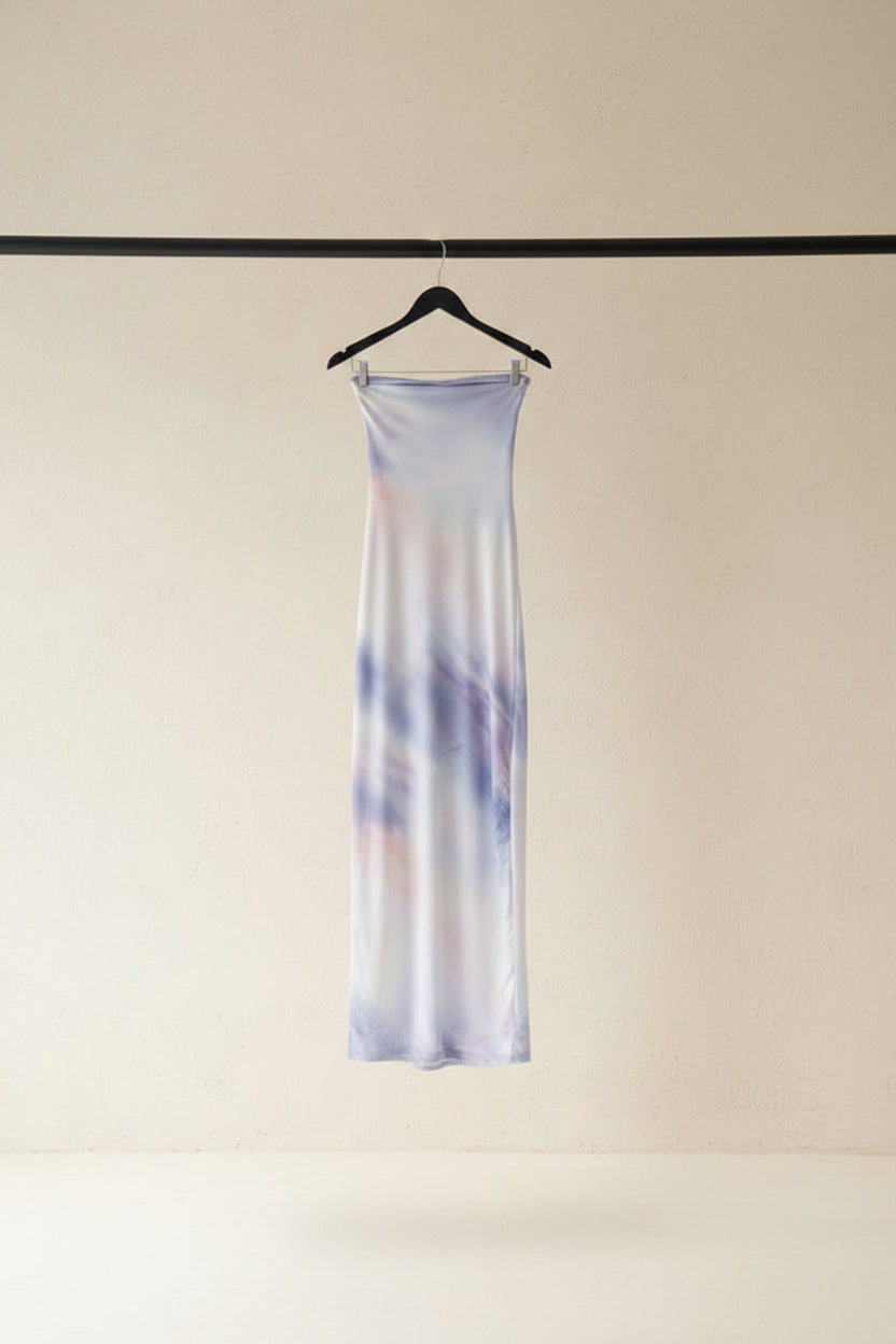 A long, sheer fabric panel or scarf with a soft tie-dye pattern in lavender, purple, and peach, hanging from a wooden hanger outdoors between tree branches in a garden courtyard.