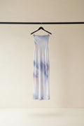 A long, sheer fabric panel or scarf with a soft tie-dye pattern in lavender, purple, and peach, hanging from a wooden hanger outdoors between tree branches in a garden courtyard.