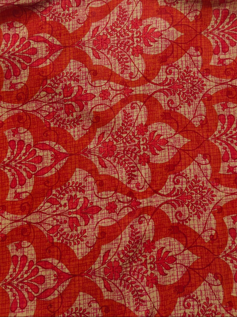 A close-up of a red and beige patterned fabric, likely a shirt or scarf, featuring a repeating ornate floral and scroll motif.
