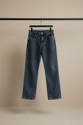 A pair of dark gray washed denim jeans hanging from a wooden hanger against a white fabric backdrop.