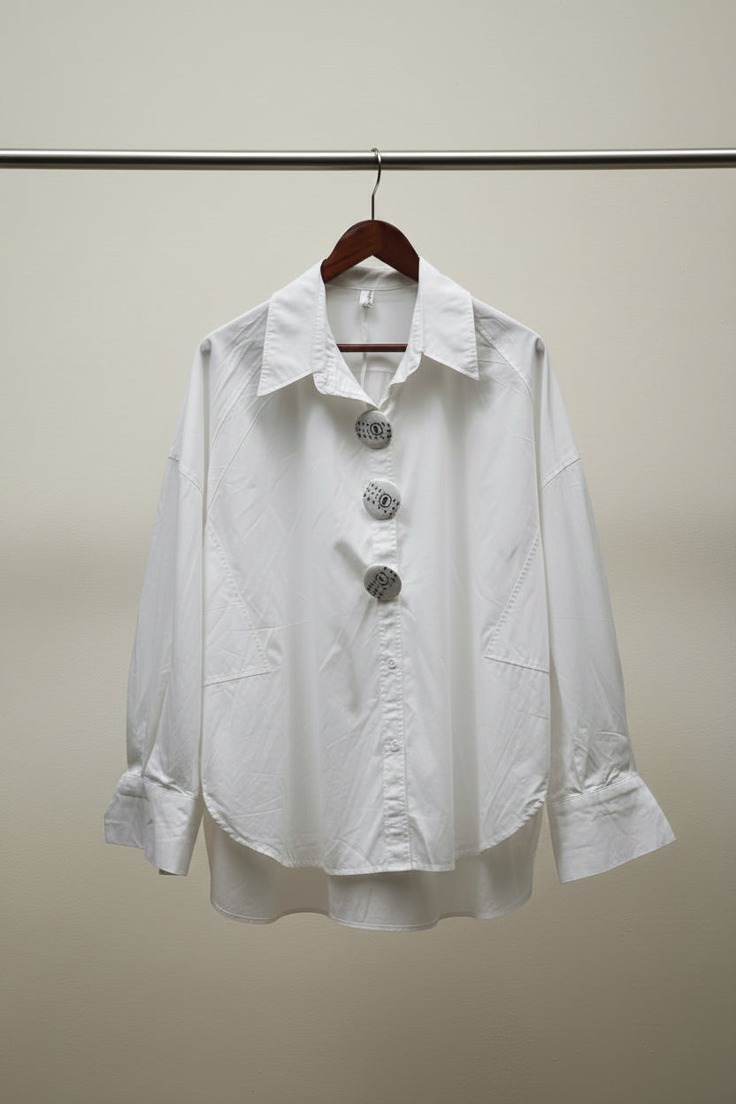 A white long-sleeve button-up shirt with three decorative silver-tone embellished buttons, hanging on a wooden hanger against an outdoor patio backdrop with branches and potted plants.