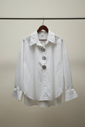 A white long-sleeve button-up shirt with three decorative silver-tone embellished buttons, hanging on a wooden hanger against an outdoor patio backdrop with branches and potted plants.