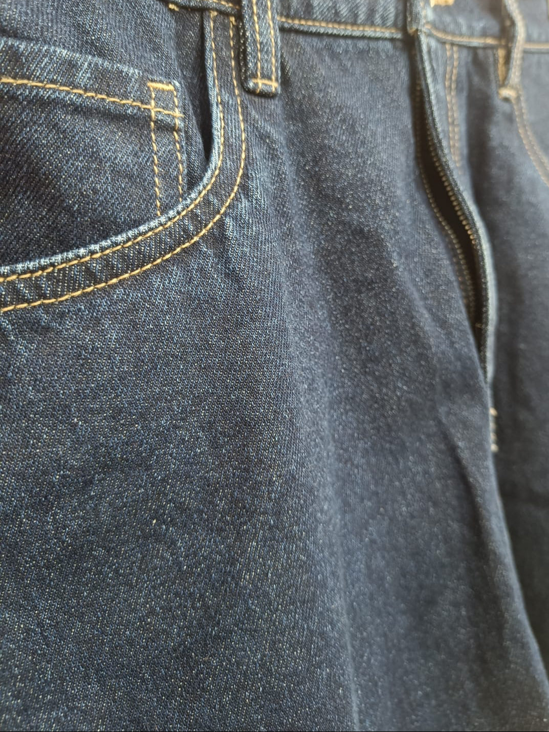 Close-up of dark blue denim jeans showing the front pocket, belt loops, and contrast yellow stitching.