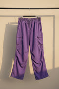 Purple cargo pants with an elastic drawstring waist and multiple utility pockets, hanging against a gray fabric backdrop.